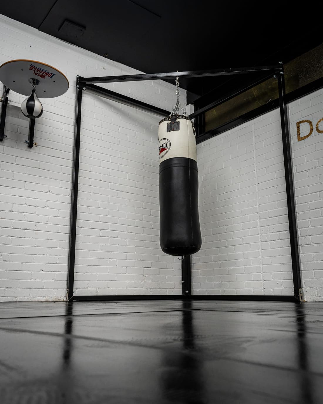 A heavy punch bag hanging from a floor-standing frame, with a speedball platform mounted on the white brick wall beside it