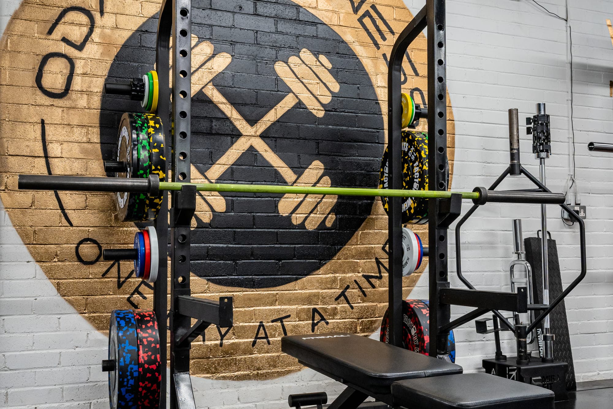 A loaded half rack and adjustable bench in front of the ODAAT Fitness roundel — a large gold-and-black crossed-dumbbell emblem painted on the brick wall, ringed with the words One Day At A Time