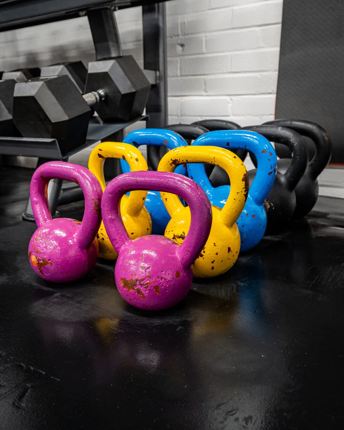 Pink, blue and yellow competition kettlebells grouped on the floor beside a rack of hex dumbbells