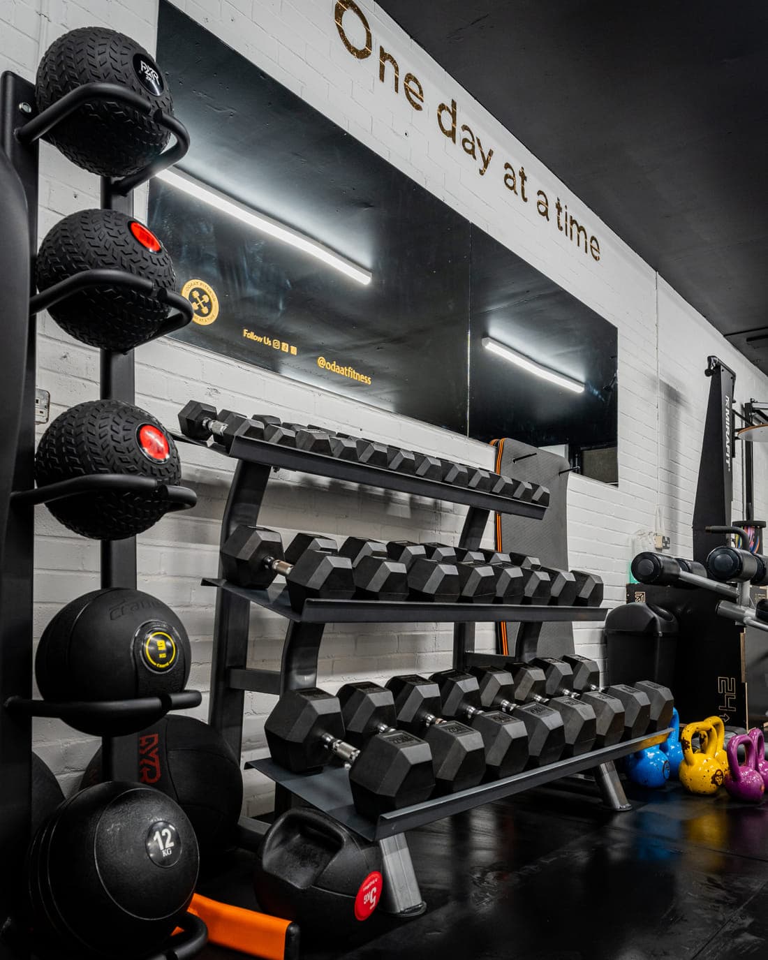 A rack of weighted medicine balls and slam balls beside a dumbbell rack, under the words One day at a time painted on the wall