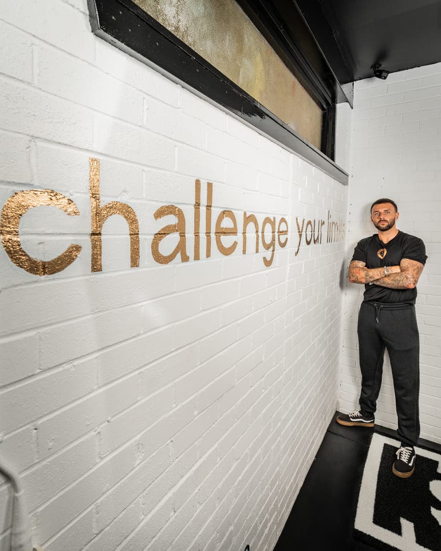 Ryan, founder of ODAAT Fitness, standing arms folded beside the words Challenge your limits painted in gold on the white brick wall