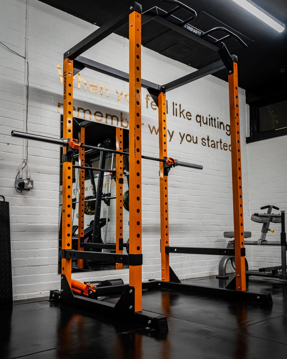 An orange and black power rack with a loaded barbell and pull-up bar, set against white brick painted with the words Remember why you started