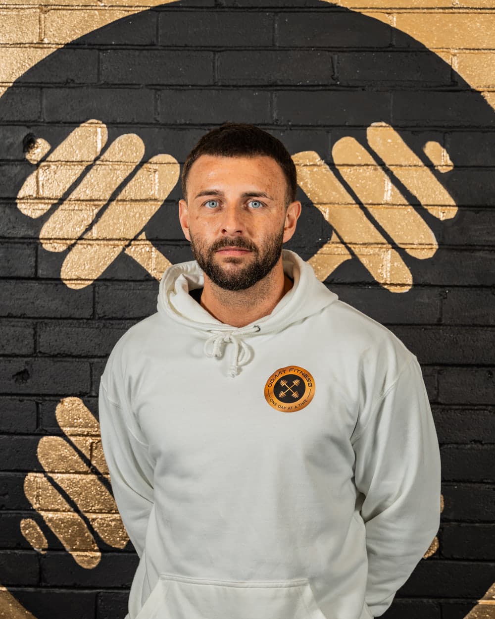 Ryan, founder of ODAAT Fitness, in a cream ODAAT Fitness hoodie, standing in front of the gold and black crossed-dumbbell roundel painted on the gym wall