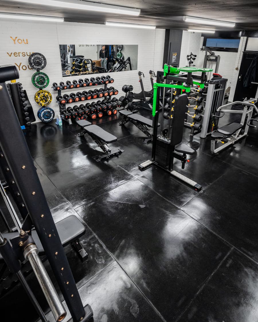 An empty, uncrowded stretch of the training floor with free benches, a plate-loaded press and racked dumbbells along the wall