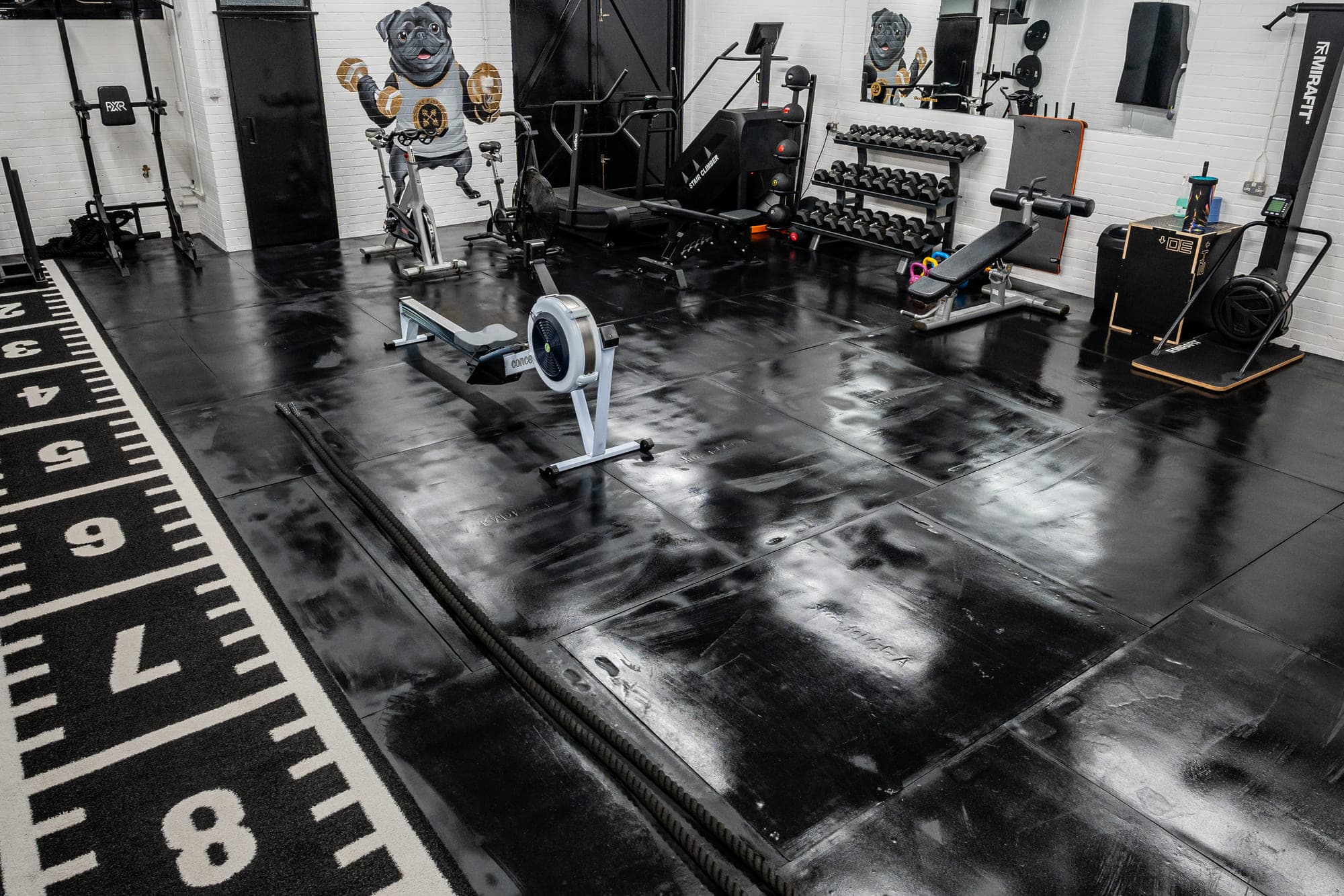 The main training floor at ODAAT Fitness, with a Concept2 rower, dumbbell rack and benches on black rubber flooring beside a marked sled track