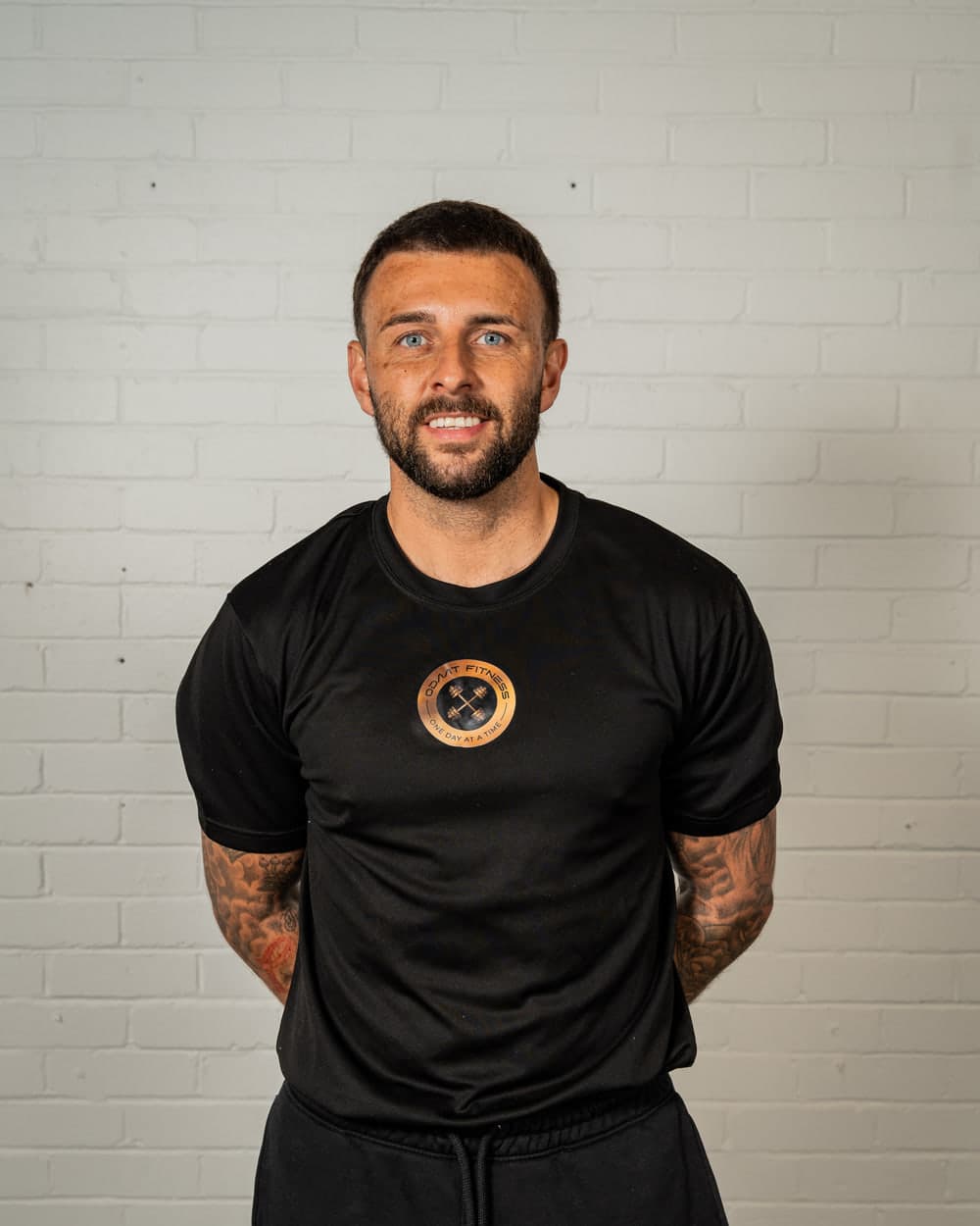 Ryan, founder of ODAAT Fitness, in a black ODAAT Fitness training top with the gold roundel badge on the chest, standing against the gym's white brick wall